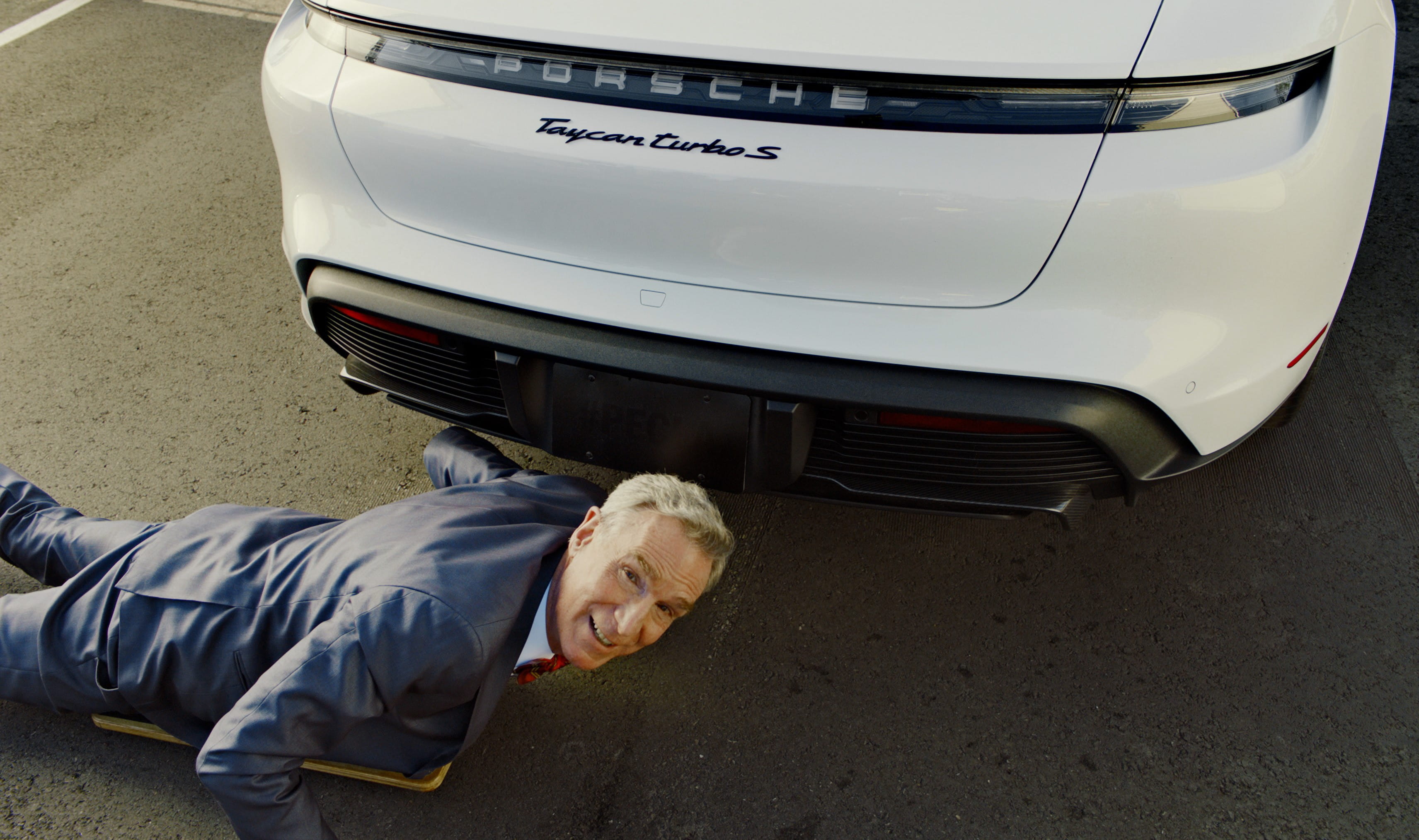 Bill Nye. Blue suit. Porsche Taycan. Under car.
