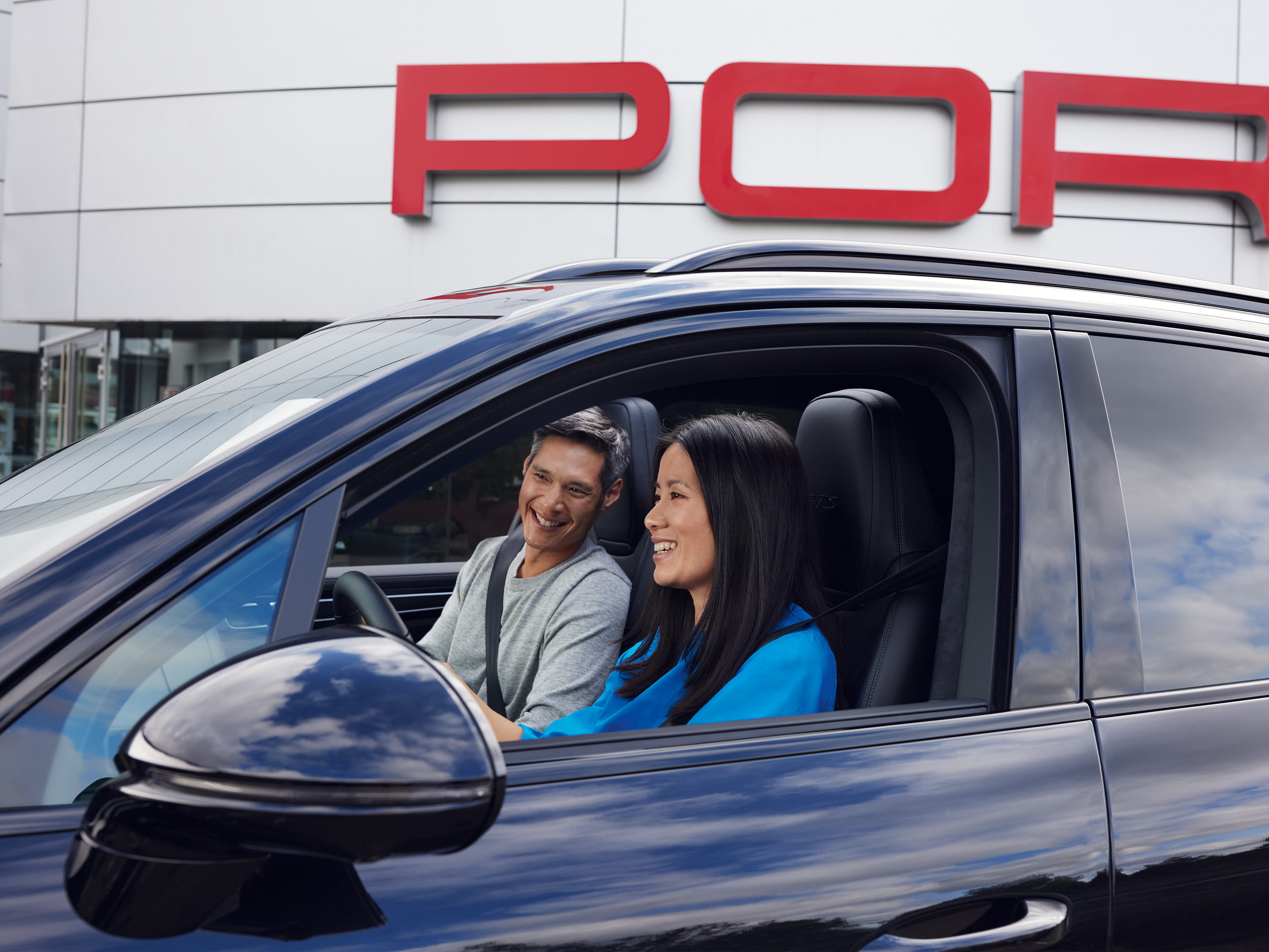 Man and woman in car, outside car dealer. Porsche Centre in background.